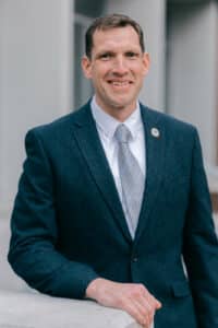 THF Spring 2024 Headshot Proofs A man in a dark blue blazer and a light blue tie stands outdoors, leaning on a stone railing. He smiles confidently and wears a pin on his left lapel, suggesting he holds a position of authority. The background features a modern building.